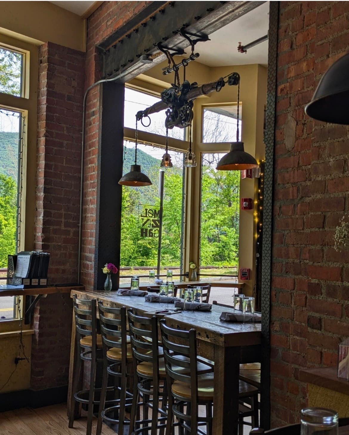 A cozy restaurant interior featuring a long wooden table, high stools, and large windows with views of greenery outside.