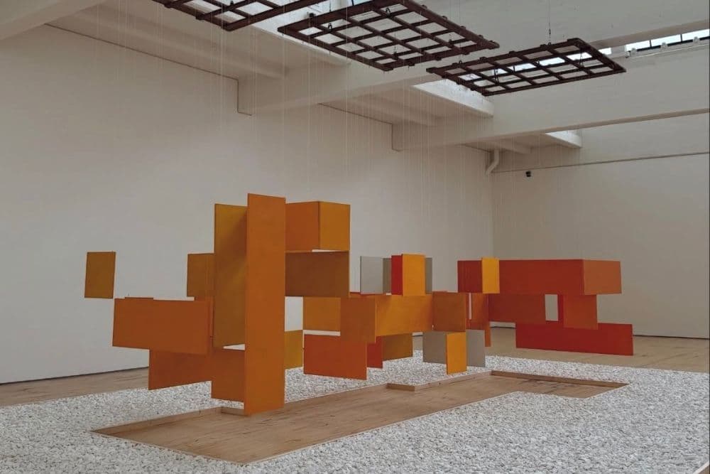 A modern abstract sculpture composed of interlocking orange and yellow geometric shapes suspended in a gallery space.