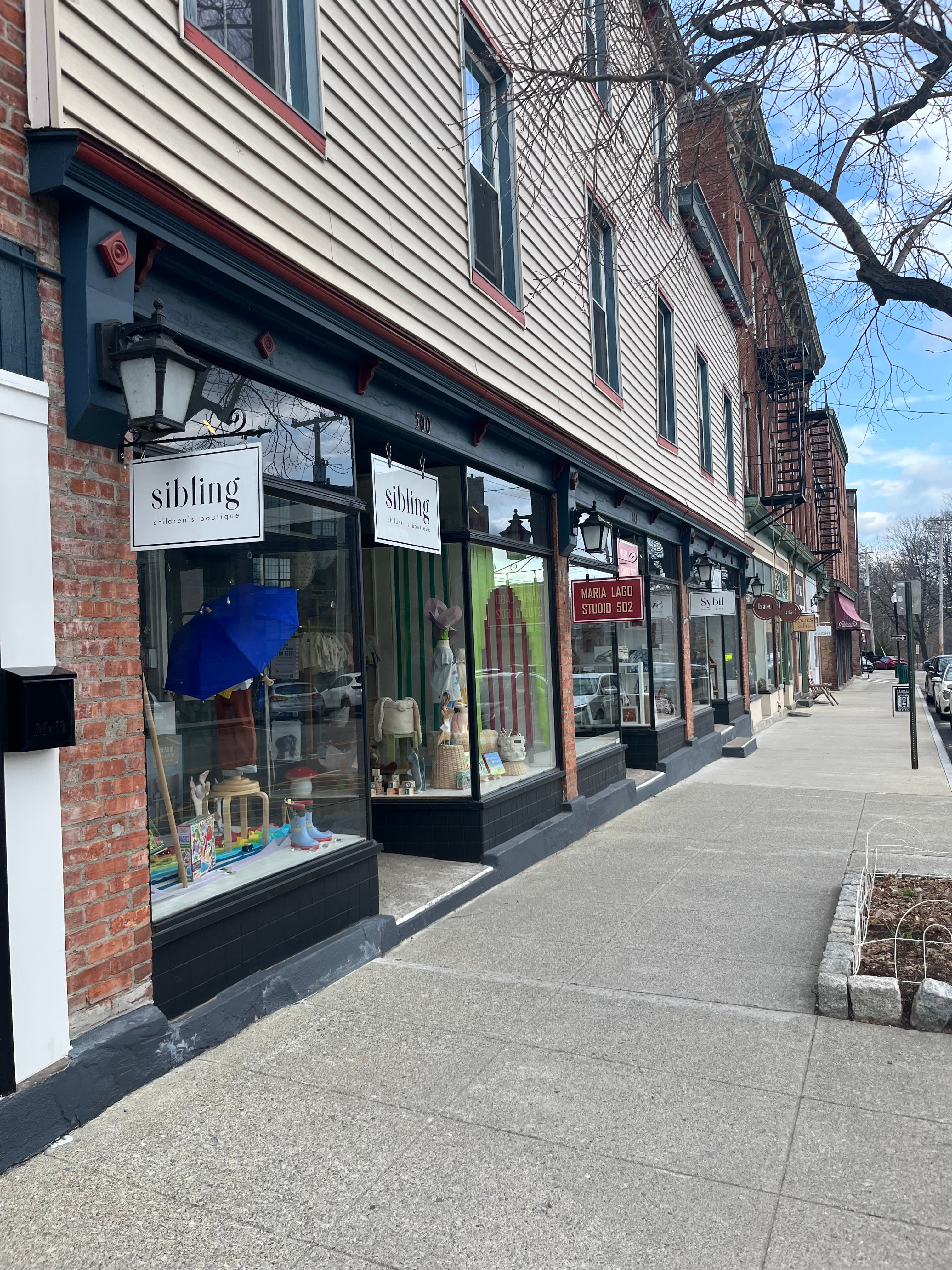 A row of storefronts, including a toy shop named "sibling," line a sidewalk on a sunny day.