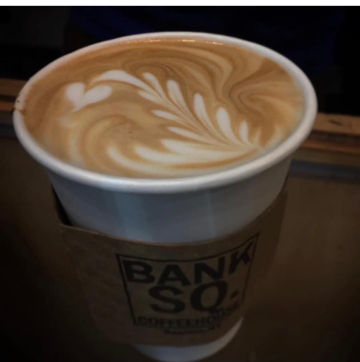 A latte with intricate heart-shaped latte art on top, served in a branded paper cup.