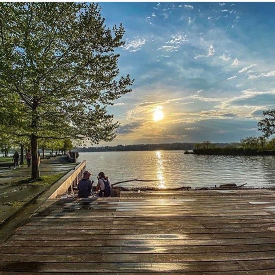 A serene lakeside scene features two people sitting on a wooden dock as the sun sets behind them.