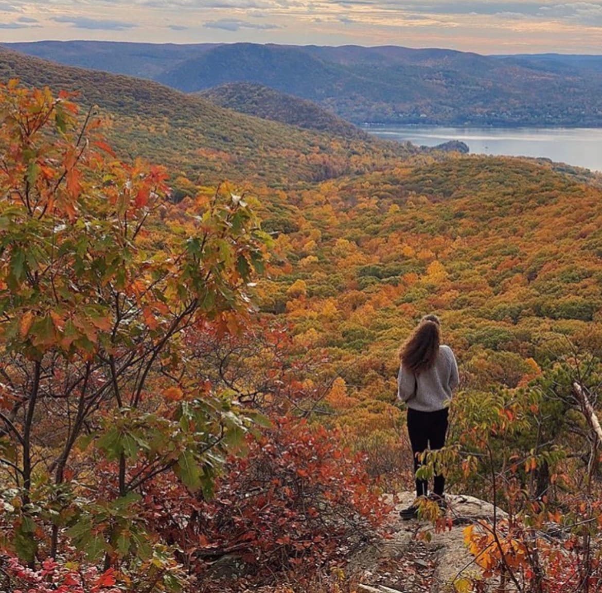 Person standing on a rocky ledge, overlooking a valley filled with vibrant autumn foliage and a river in the distance.