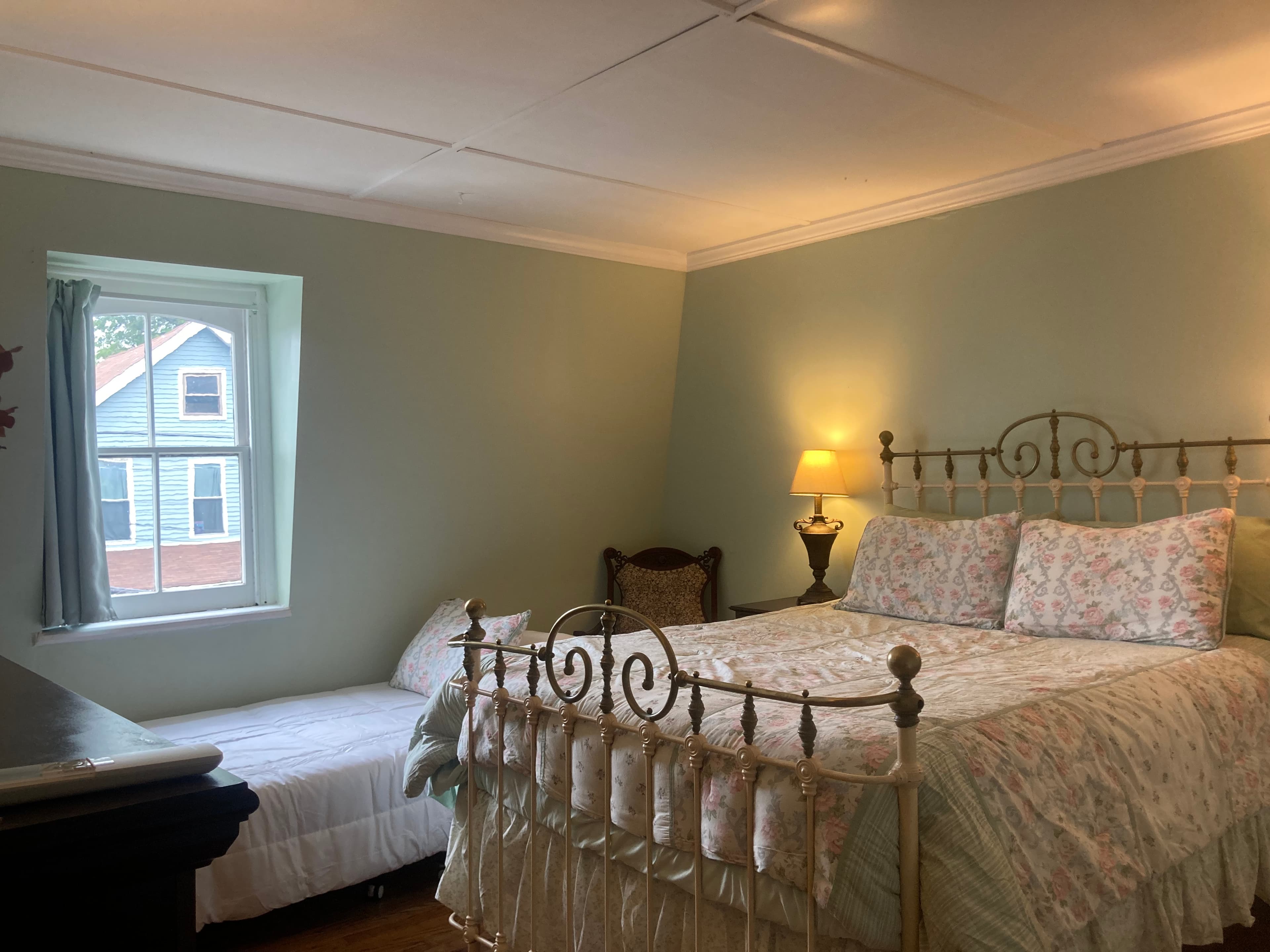 A cozy bedroom with a floral-patterned bed, an armchair, and a window revealing a glimpse of an exterior house.