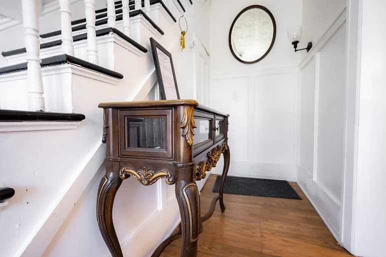 A wooden console table stands in a bright hallway, with stairs leading up and a round mirror on the wall.